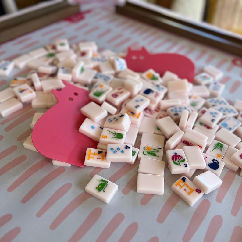 Peut inclure: Un ensemble de tuiles de Mahjong ivoire, avec des symboles et des chiffres color&eacute;s, dispers&eacute;es sur une surface bleu clair avec une d&eacute;coupe rose en forme de chat. Les tuiles pr&eacute;sentent divers motifs, dont des fleurs, du bambou et des points. Un jeu de soci&eacute;t&eacute; classique.