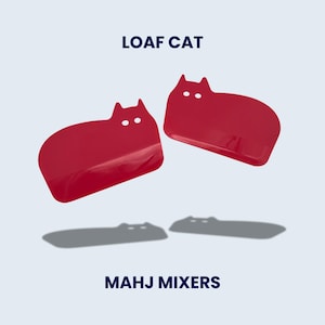 May include: Two red cat-shaped kitchen tools with the text "LOAF CAT" above them and "MAHJ MIXERS" below. The tools have a rounded shape with a cat head design. The tools are casting a shadow on a light blue background.
