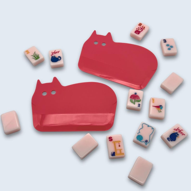 Peut inclure: Deux plateaux rouges en forme de chat et plusieurs tuiles de Mahjong sont dispers&eacute;s sur une surface bleu clair. Les tuiles sont blanches avec des illustrations et des chiffres color&eacute;s. Les plateaux ont deux d&eacute;coupes circulaires pour les yeux.