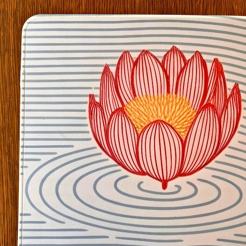 以下が含まれることがあります： 赤い蓮の花と黄色の中心、詳細な線が特徴の長方形のマウスパッド。 デザインは、水色の水平ストライプと同心円の背景に配置され、穏やかなデザインを作り出しています。