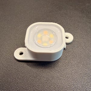 May include: A white, square-shaped LED light fixture with a clear, circular lens. The lens reveals several small, yellow-orange lights. The fixture has two small mounting tabs with holes for screws.