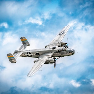 May include: A silver vintage airplane with a star emblem on the wing and tail, flying against a cloudy blue sky. The plane has two propellers and a glass cockpit. The image is signed by JL 4 Photo.