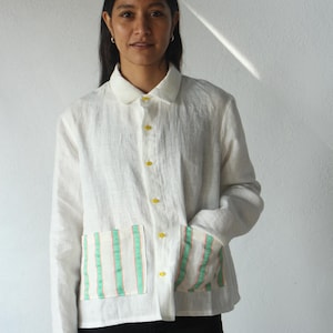 May include: A white linen jacket with long sleeves, a collar, and yellow buttons. The jacket features two front pockets with vertical stripes in mint green and coral. The person is wearing black pants.