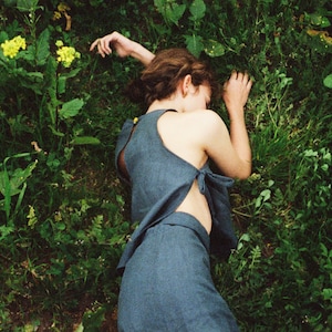 Op de afbeelding: Een vrouw in een blauwe linnen jurk met een lage rug en een strik in de taille ligt op haar zij in een veld met groen gras en gele bloemen.