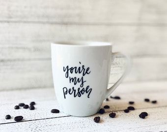 May include: White ceramic mug with the text "you're my person" in dark blue script. Scattered coffee beans surround the mug. The mug has a curved handle and is set against a white wooden background.