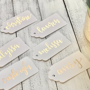 May include: Six rectangular frosted translucent tags with rounded corners and a hole punch at one end. Each tag features a name written in metallic gold cursive script. The names displayed are christine, lauren, alyssa, melissa, carleigh, and avery.