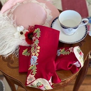 May include: A pink hat with a white feather trim, red and white floral embroidered red gloves, and a white teacup with a blue and gold rim on a wooden table.