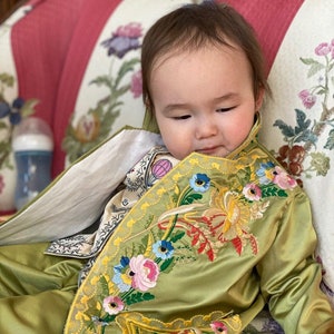 Puede incluir: Un bebé con una chaqueta de satén verde con bordado floral de colores. La chaqueta tiene un ribete amarillo y se lleva sobre una camisa blanca con estampado floral.
