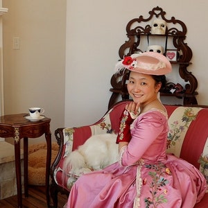 May include: A woman wearing a pink dress and a pink hat with a red flower sits on a striped couch with a white dog. The couch has floral patterns and is in a room with a fireplace and a decorative shelf.