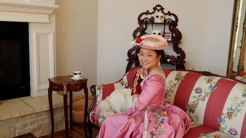 May include: A woman wearing a pink dress and a pink hat with a red flower sits on a striped couch with a white dog. The couch has floral patterns and is in a room with a fireplace and a decorative shelf.