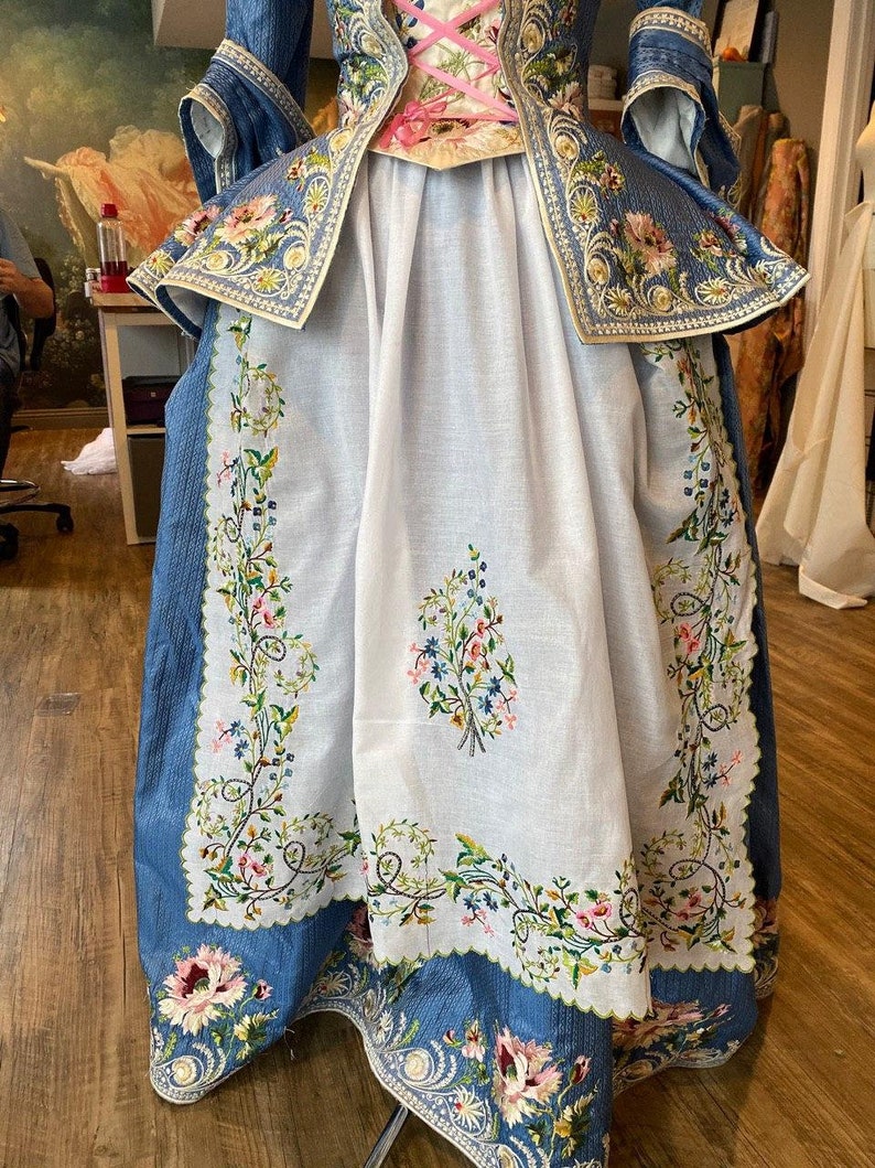 May include: A white skirt with blue floral embroidery around the hem. The skirt is layered over a blue skirt with a floral pattern and gold trim.