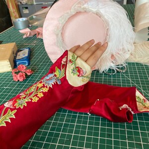 May include: A red satin sleeve with floral embroidery and a white trim. The sleeve is pinned to a green cutting mat.