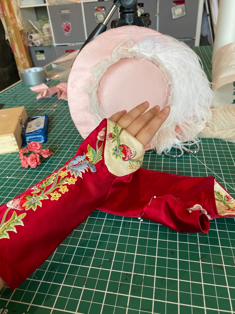 May include: A red satin sleeve with floral embroidery and a white trim. The sleeve is pinned to a green cutting mat.