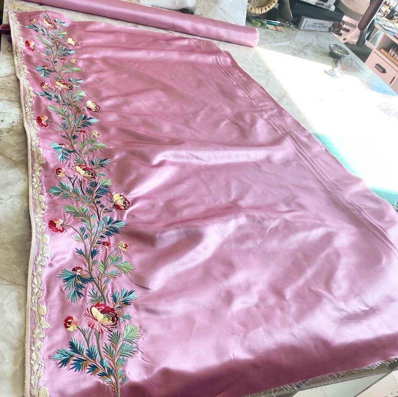 May include: Pink satin fabric with a floral embroidery design along the edge. The embroidery features pink, green, and white flowers.