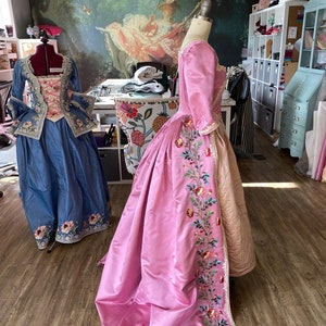 May include: Two vintage-style dresses on dress forms. The dress on the left is blue with white embroidery. The dress on the right is pink with floral embroidery.