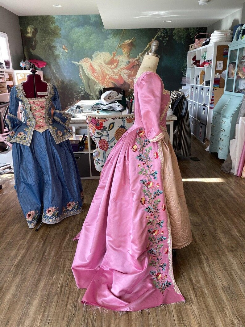 May include: Two vintage-style dresses on dress forms. The dress on the left is blue with white embroidery. The dress on the right is pink with floral embroidery.