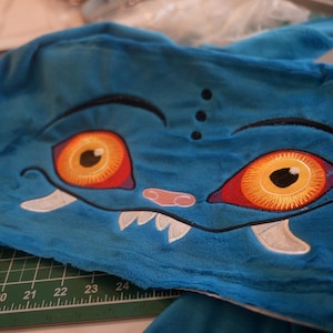 May include: A blue plush fabric with an embroidered monster face. The face features large orange and red eyes, white fangs, and a pink nose. The design is detailed with black outlines and stitching.