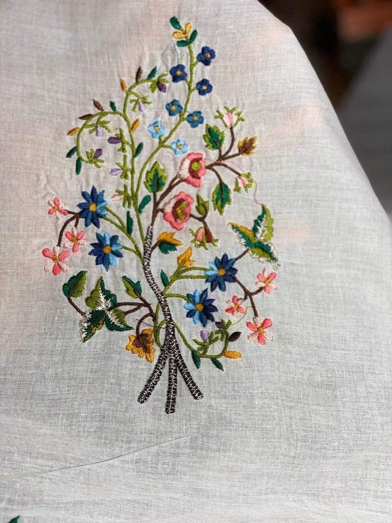May include: White fabric with embroidered floral design featuring pink, blue, and yellow flowers with green leaves.