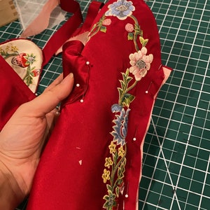 May include: A red fabric piece with a floral embroidery design in shades of pink, blue, green, and yellow. The fabric is pinned to a green cutting mat.