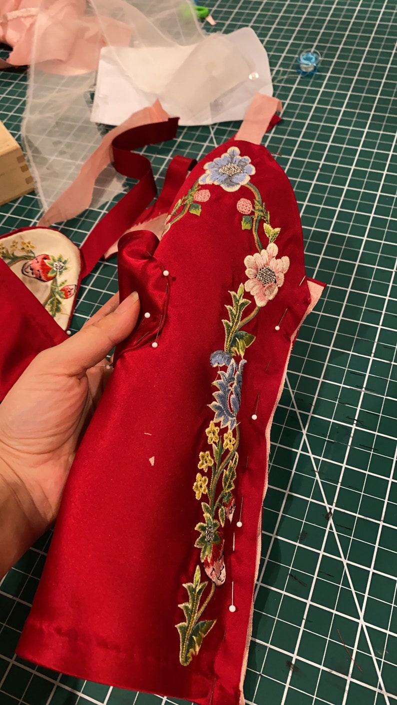 May include: A red fabric piece with a floral embroidery design in shades of pink, blue, green, and yellow. The fabric is pinned to a green cutting mat.