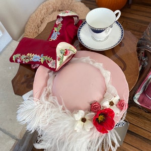 May include: A pink hat with white feathers and red and white flowers. The hat is sitting on a table with a red embroidered glove and a teacup and saucer.