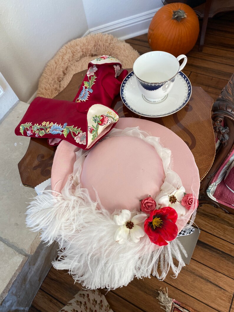 May include: A pink hat with white feathers and red and white flowers. The hat is sitting on a table with a red embroidered glove and a teacup and saucer.