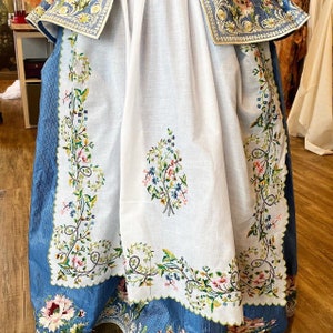 May include: A white tablecloth with blue embroidered floral designs. The tablecloth has a scalloped edge and is made of a delicate fabric.