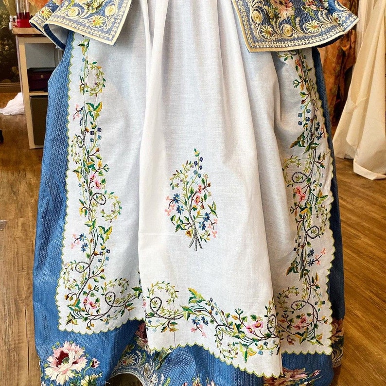 May include: A white tablecloth with blue embroidered floral designs. The tablecloth has a scalloped edge and is made of a delicate fabric.
