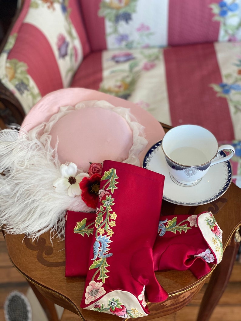 May include: A pink hat with a white feather trim, red and white floral embroidered red gloves, and a white teacup with a blue and gold rim on a wooden table.