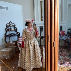 May include: A woman wearing a long, cream-colored dress with a floral pattern. She is wearing a pink hat with a feather and red gloves. She is standing in front of a mirror and taking a selfie.