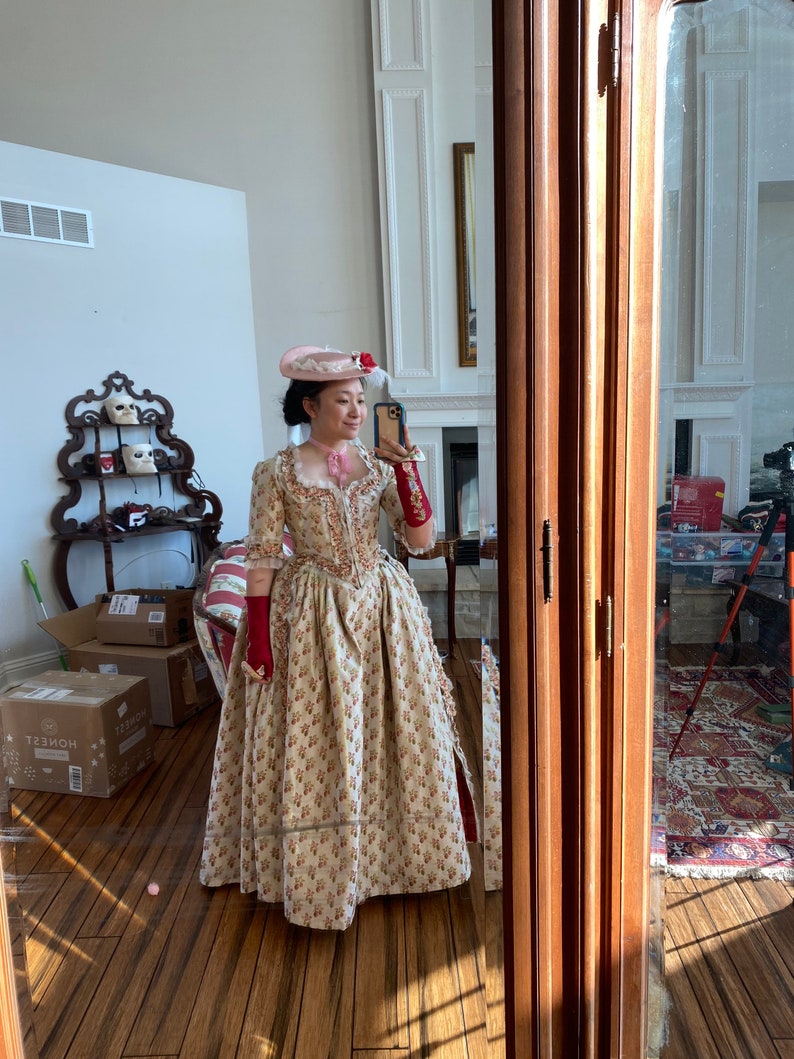May include: A woman wearing a long, cream-colored dress with a floral pattern. She is wearing a pink hat with a feather and red gloves. She is standing in front of a mirror and taking a selfie.