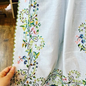 May include: A white linen tablecloth with a delicate floral embroidery design. The embroidery features green leaves, pink, blue, and yellow flowers.