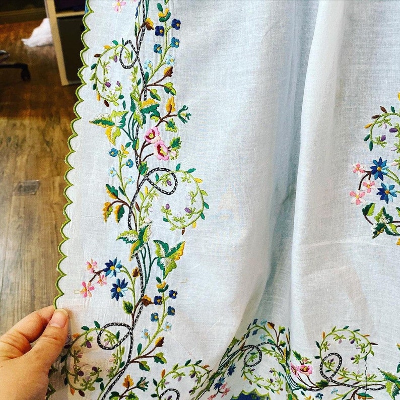 May include: A white linen tablecloth with a delicate floral embroidery design. The embroidery features green leaves, pink, blue, and yellow flowers.