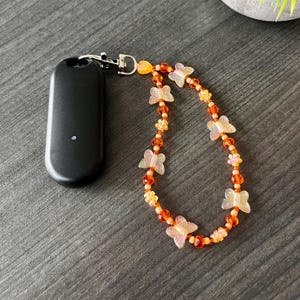 Może przedstawiać: Czarne etui na telefon z przyczepionym do niego koralikowym wisiorkiem w kształcie pomarańczowego i białego motyla.