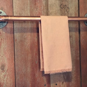 May include: A copper pipe towel rack with industrial style flanges mounted on a wooden wall. A light peach colored towel is hanging on the rack.