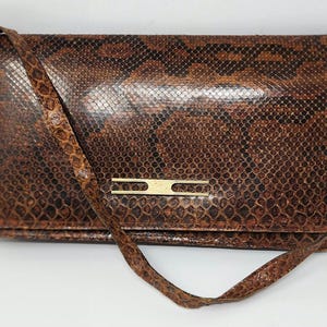 May include: A brown and black snakeskin-patterned clutch with a matching shoulder strap. The rectangular bag features a gold-colored clasp and a flap closure. The bag is made of a textured material.