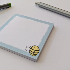 May include: A blue notepad with a white centre and a cartoon bee illustration on the bottom right corner. The notepad is on a white surface.