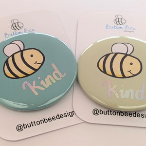 May include: Two circular badges featuring a cartoon bee. One is light blue, the other is pale green. The word "Kind" is written on each in a holographic font. They are displayed on white cards with the Button Bee Designs logo.