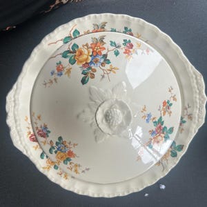 May include: Cream-coloured serving dish with a lid, featuring a floral pattern in orange, yellow, blue, and pink. The dish has a scalloped edge and a raised floral design on the lid. Ideal for serving food.