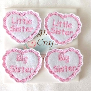 May include: Four pink and white heart-shaped fabric patches with embroidered text. Two patches say "Little Sister" and two patches say "Big Sister".