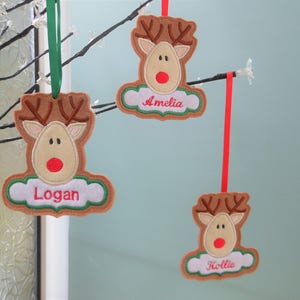 K&ouml;nnte beinhalten: Drei personalisierte Rentier-Weihnachtsbaumschmuck. Jedes Ornament zeigt einen braunen Rentierkopf mit Geweih, einer roten Nase und einem Namen auf einer wei&szlig;en, wolkenf&ouml;rmigen Basis. Sie h&auml;ngen an gr&uuml;nen und roten B&auml;ndern.