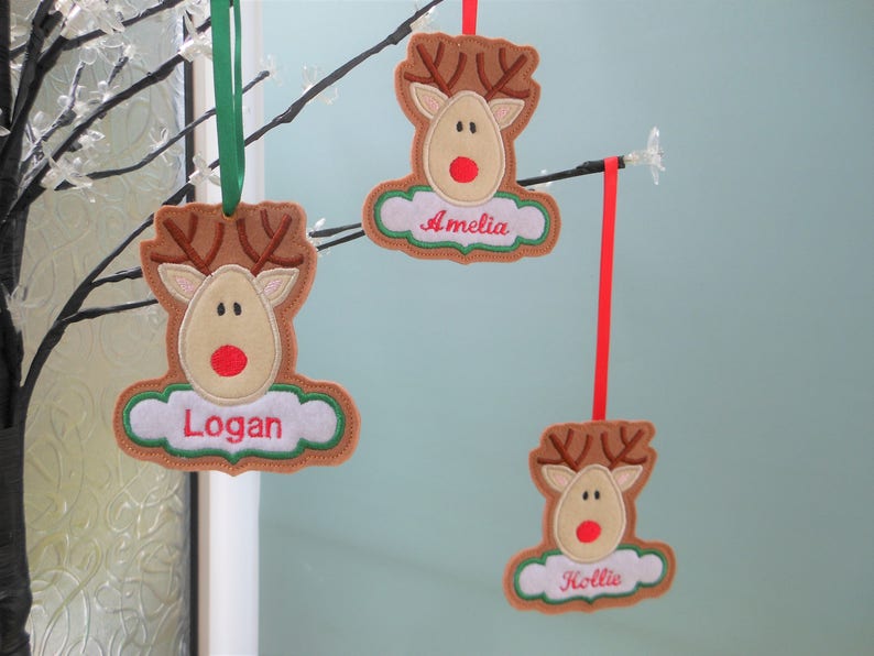 K&ouml;nnte beinhalten: Drei personalisierte Rentier-Weihnachtsbaumschmuck. Jedes Ornament zeigt einen braunen Rentierkopf mit Geweih, einer roten Nase und einem Namen auf einer wei&szlig;en, wolkenf&ouml;rmigen Basis. Sie h&auml;ngen an gr&uuml;nen und roten B&auml;ndern.