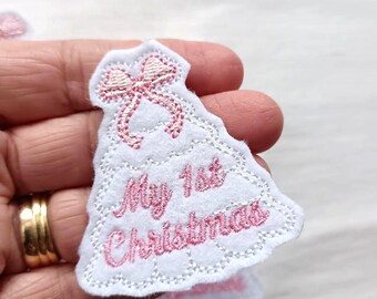 May include: A white felt patch shaped like a Christmas tree with scalloped edges. The patch features pink embroidered stitching, a pink bow at the top, and the pink embroidered text "My 1st Christmas" centered on the front surface.