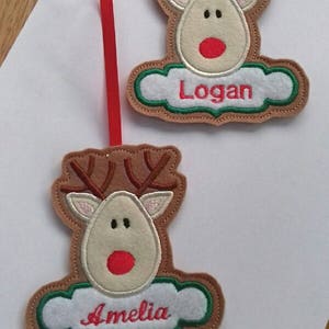 K&ouml;nnte beinhalten: Zwei Rentier-Ornamente mit braunen Geweihen, beigefarbenen Gesichtern und roten Nasen. Jedes Ornament hat ein wei&szlig;es, wolkenf&ouml;rmiges Banner mit einem rot gestickten Namen. Eines tr&auml;gt "Logan" und das andere "Amelia". Ein rotes Band ist befestigt.