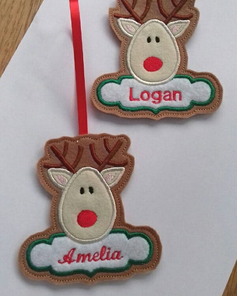 K&ouml;nnte beinhalten: Zwei Rentier-Ornamente mit braunen Geweihen, beigefarbenen Gesichtern und roten Nasen. Jedes Ornament hat ein wei&szlig;es, wolkenf&ouml;rmiges Banner mit einem rot gestickten Namen. Eines tr&auml;gt "Logan" und das andere "Amelia". Ein rotes Band ist befestigt.