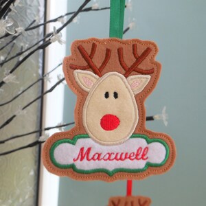 K&ouml;nnte beinhalten: Ein Weihnachtsornament aus Filz in Form eines Rentiers mit braunem Geweih, einem beigen Gesicht und einer roten Nase. Der Name "Maxwell" ist in roter Schrift auf einem wei&szlig;en Banner gestickt. Ein gr&uuml;nes Band ist zum Aufh&auml;ngen befestigt.