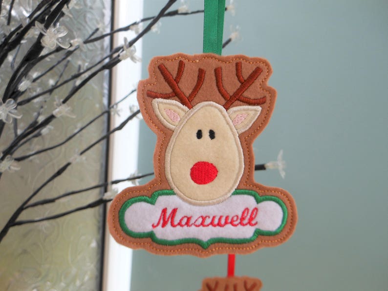 K&ouml;nnte beinhalten: Ein Weihnachtsornament aus Filz in Form eines Rentiers mit braunem Geweih, einem beigen Gesicht und einer roten Nase. Der Name "Maxwell" ist in roter Schrift auf einem wei&szlig;en Banner gestickt. Ein gr&uuml;nes Band ist zum Aufh&auml;ngen befestigt.