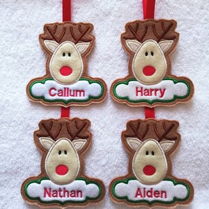 K&ouml;nnte beinhalten: Vier Weihnachtsbaumschmuck aus Filz in Form von Rentieren, jeweils mit einem anderen Namen: Callum, Harry, Nathan und Aiden. Die Rentiere haben braune Geweihe, beige Gesichter und rote Nasen. Jeder Schmuck h&auml;ngt an einem roten Band.