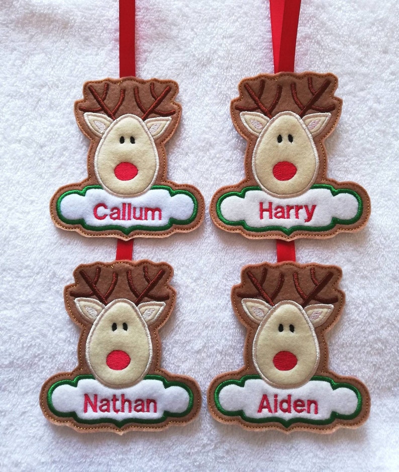 K&ouml;nnte beinhalten: Vier Weihnachtsbaumschmuck aus Filz in Form von Rentieren, jeweils mit einem anderen Namen: Callum, Harry, Nathan und Aiden. Die Rentiere haben braune Geweihe, beige Gesichter und rote Nasen. Jeder Schmuck h&auml;ngt an einem roten Band.
