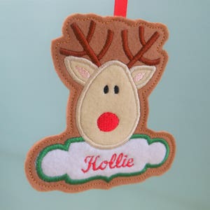 K&ouml;nnte beinhalten: Ein Weihnachtsornament aus Filz in Form eines Rentiers. Es hat braune H&ouml;rner, ein beiges Gesicht und eine rote Nase. Der Name "Hollie" ist rot auf einem wei&szlig;en, wolkenf&ouml;rmigen Bereich gestickt.